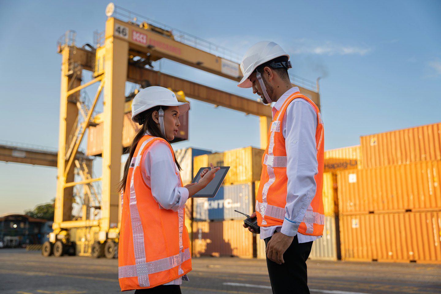Two logistics workers discuss port operations with containers and cranes.