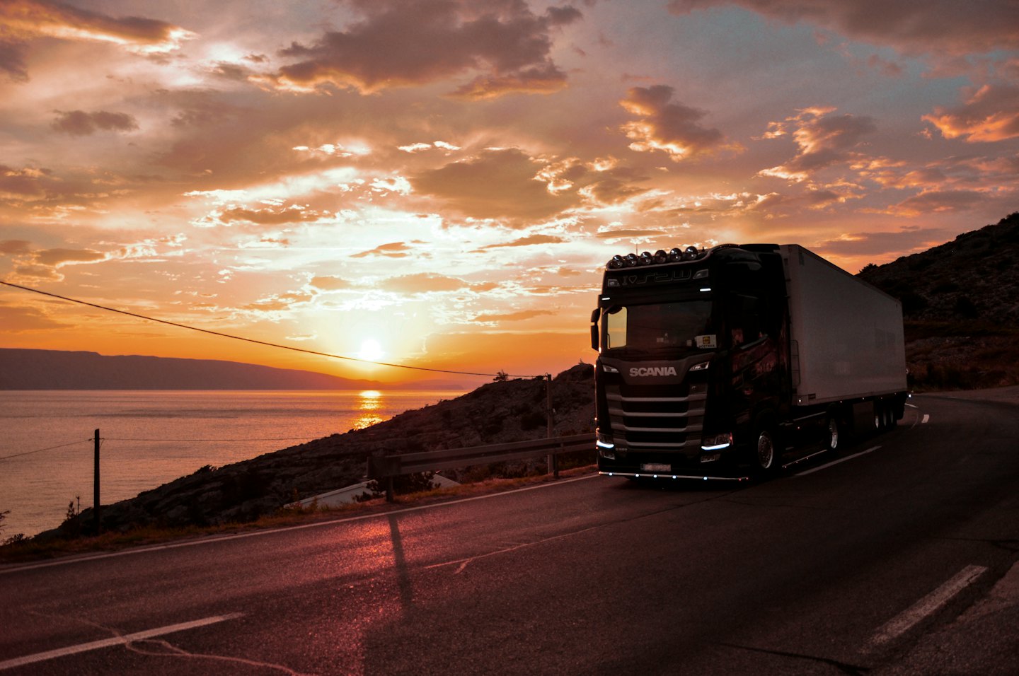 Black semi-truck drives on coastal road at sunset.