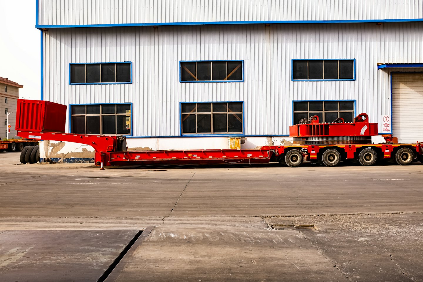 A red lowboy trailer parked beside an industrial building.